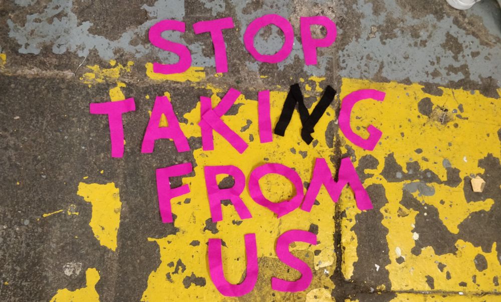 fuschia-coloured felt cut-outs of letters arranged to say "STOP TAKING FROM US" in capital letters. the 'n' is in black felt. they lay over a concrete ground that has some chipped grey and yellow paint.
