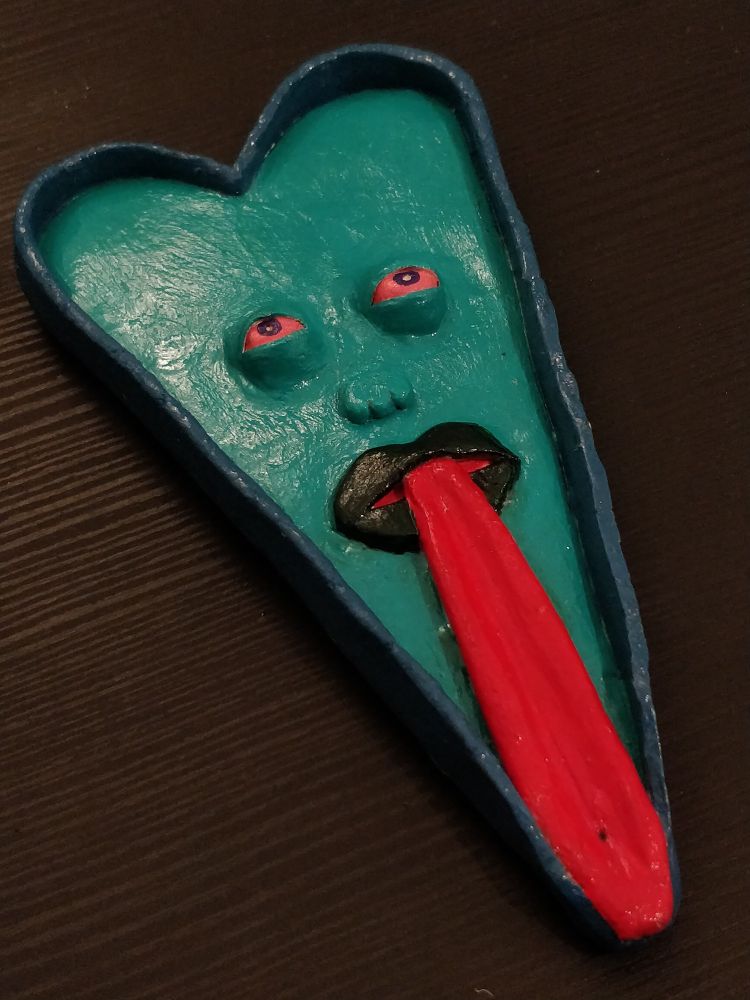 a clay ashtray that's love heart shaped. the heart has eyes, which are squinting and bloodshot, a small nose, and a mouth with a long tongue sticking out. the tongue is curved inwards so a cigarette or joint can lay on it.