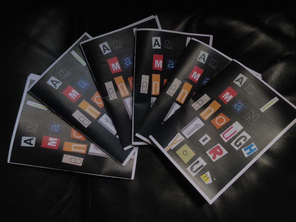 six copies of a zine titled "am i man enough for you?!" laid out in an arch together against a black pleather sofa. the zine's cover is a black background with cut-out magazine letters spelling the title.