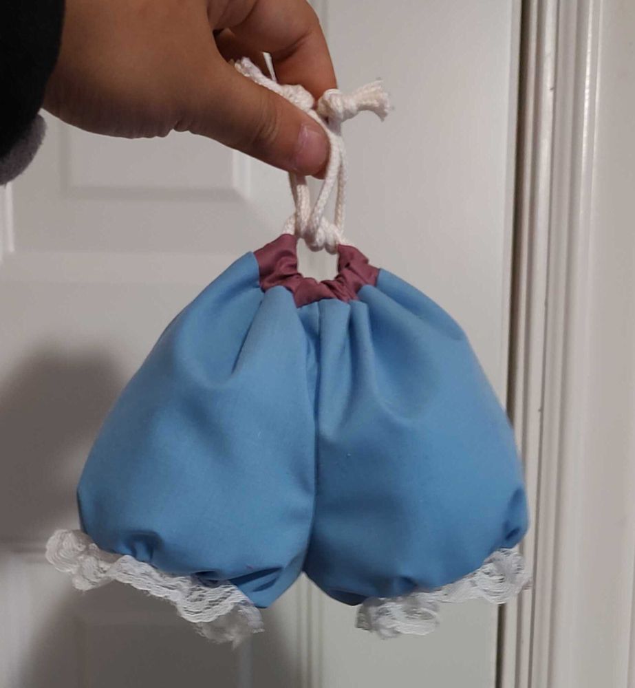 A picture of my """bag""" prototype dangling by the cinched up drawstring. Instead of the printed stripes and appliqued hearts of the real article, this prototype has a plain blue fabric body. Lace was used at the bottoms instead of ruffled minky. Sure looks scrotal, doesn't it.
