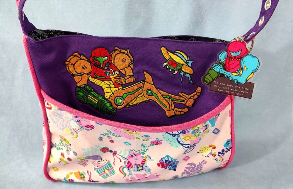 A shoulder bag I made with Kirby fabric and an embroidered Samus leaning back, enjoying a tropical drink. A Metroid in a sunhat is hanging out with her. And I got a Fusion Samus keychain as an accessory. This is my current daily bag. Delightfully eclectic!