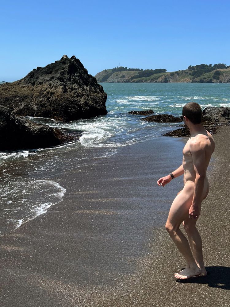 (non-sexual nudity) A nude man stands on a beach looking toward the rocks and waves.