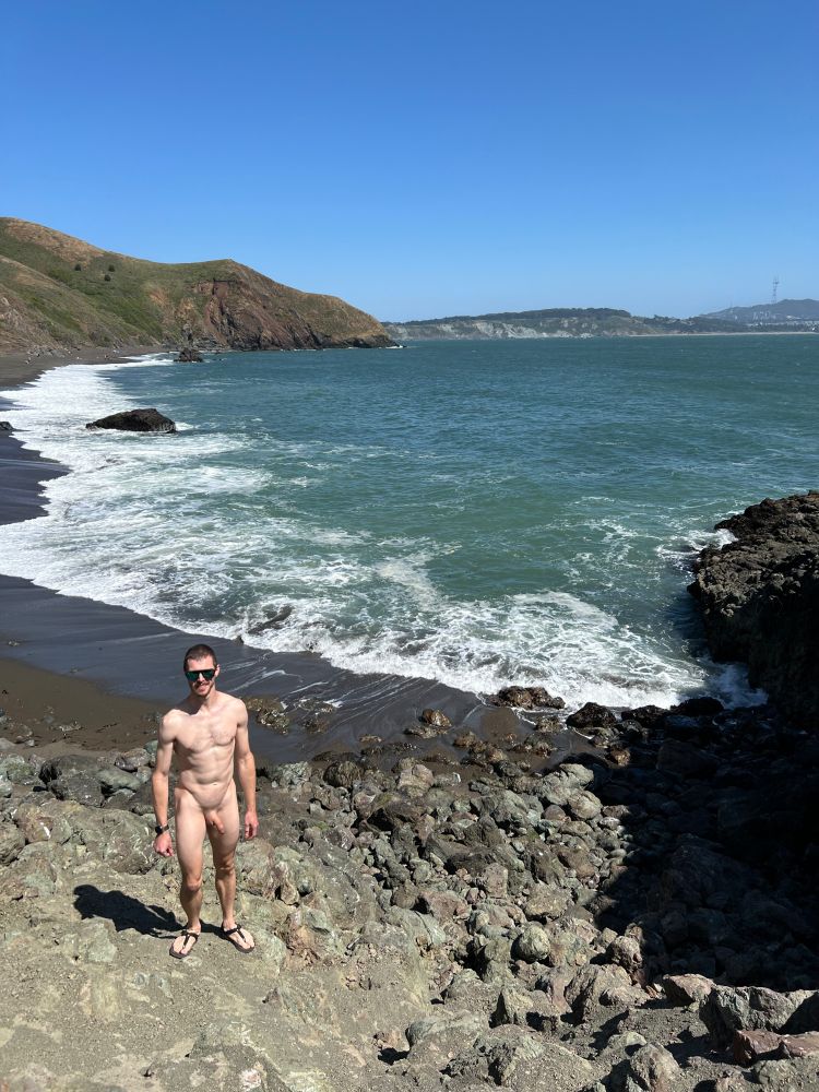 A nude man stands on rocks near an expansive ocean beach.