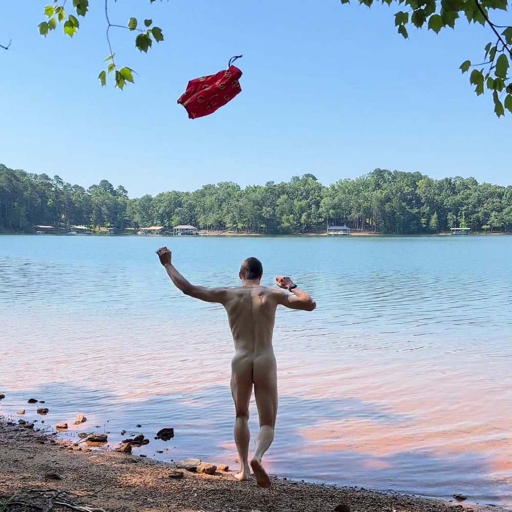 A nude man seen from behind runs into a lake. The swimsuit from the previous photo is shown thrown into the air behind him.