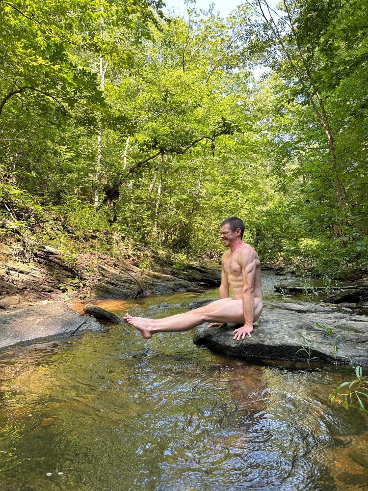 A nude man in an L sit position near a river in the woods.