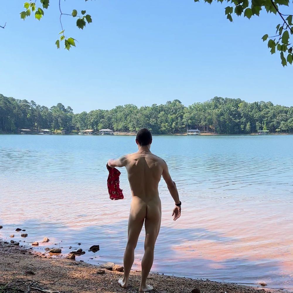 A nude man seen from behind holds a swimsuit in his left hand. He is about to throw in the air.