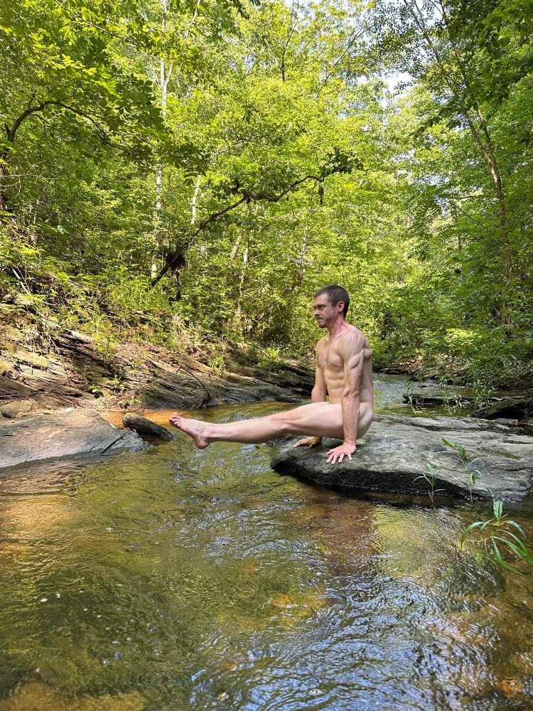 A nude man in an L sit position near a river in the woods.