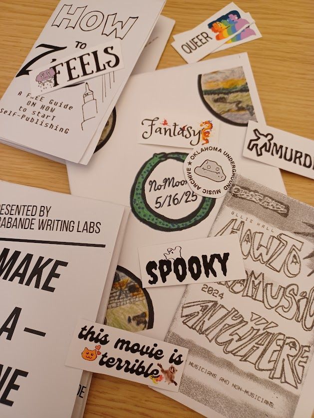 Assorted zines and stickers in an ARTFULLY ARRANGED scattering on a table. The zines are small folded titles such as "Make a Zine" though there is one special performance zine from No Moore. There is a sticker from Oklahoma Music Archive and there are some fun DVD movie spine labels such as "this movie is terrible" with a confounded raccoon on it.