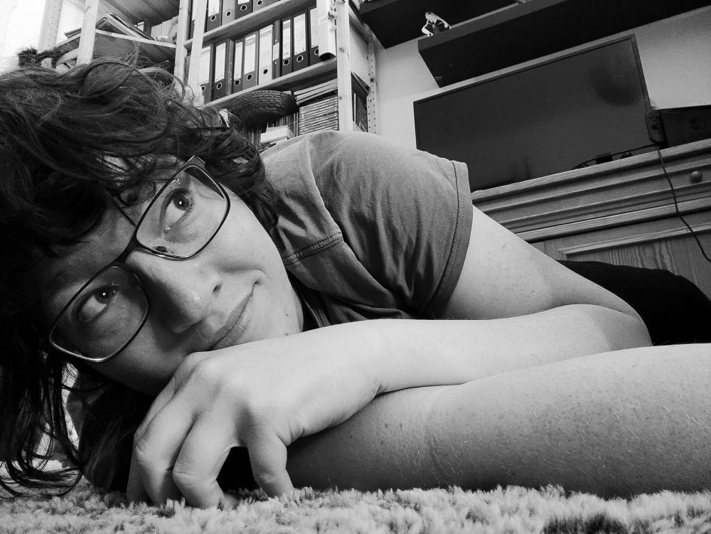 Ein Foto in schwarz-weiß von mir, auf dem Teppich liegend. Ich liege bäuchlings, ein Arm ist ausgestreckt, der andere liegt locker darüber. Lächelnd schaue ich aus dem Bild heraus.