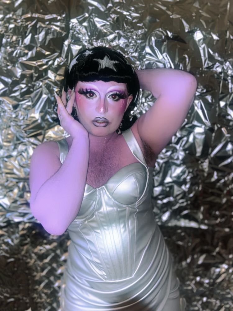 A portrait photograph of femme presenting drag artist Eden Spaghetti. They are posed elegantly against a silver crinkly background. Eden has their skin painted purple and has dramatic makeup to accentuate their features. They are wearing a black flippy bob wig with a fringe and white star decals. They are wearing a silver pleather dress with futuristic paneling