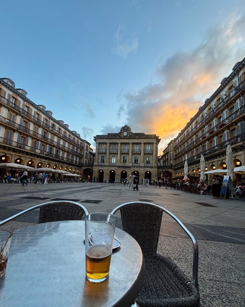 A blue sky with sunset lit clouds above a Spanish square, Constitución Plaza. There are bars all around and the town hall at the back. In the foreground is a table and a beer.