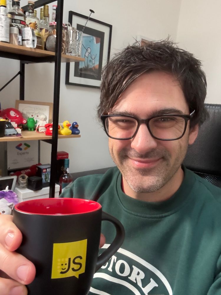 A selfie of me sitting at my desk holding a cup of coffee with the JSNation logo on it. I'm smiling at the camera and behind me are shelves with rubber ducks on.