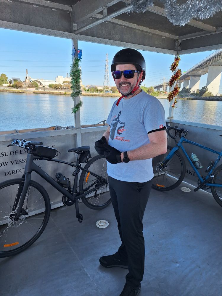 A man in a cycling helmet stands on a small boat, 2 bikes are behind him. The boat is on a river, with a large  high bridge in the background.  The sun is shining, the sky and the river are bright blue. 