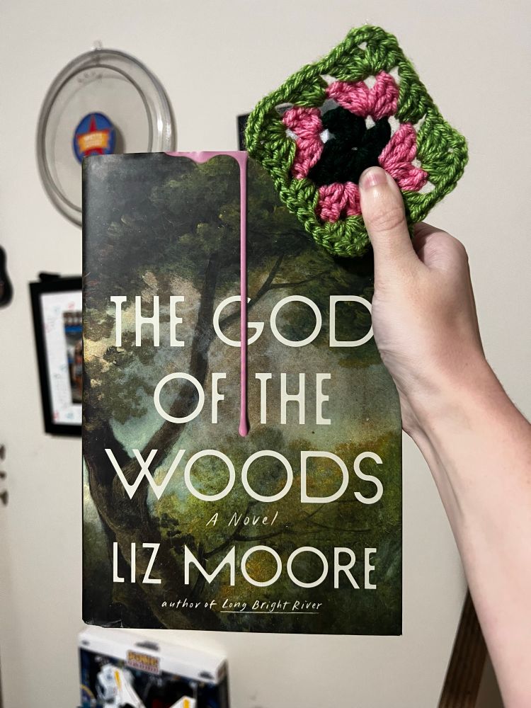 A picture of a hand holding The God of the Woods by Liz Moore and a crocheted granny square. From the center going outward, the granny square is dark green, pink, and light green to match the cover of the book.