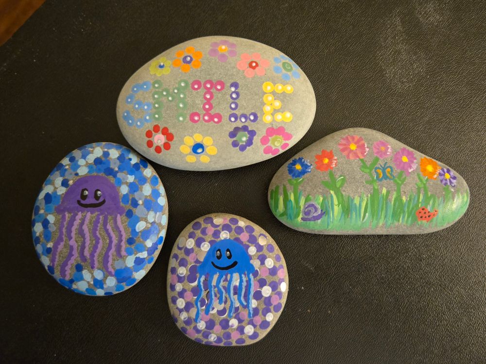 Four painted rocks. A purple jellyfish on a shades of blue dot background, a blue jellyfish on a shades of purple dot background, a little flower garden with a snail a butterfly and a ladybug, and a rock that says smile and has flowers on it.