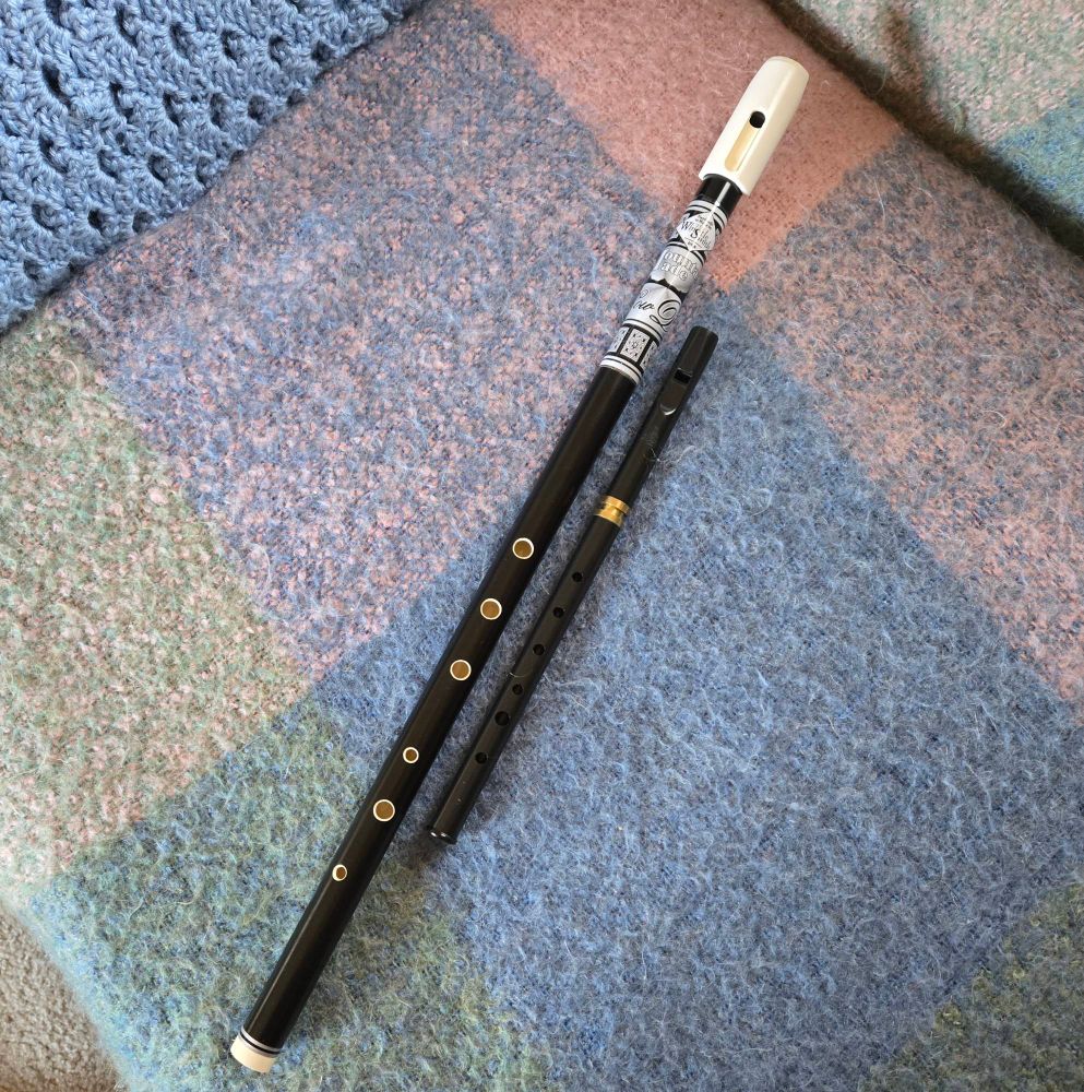 Lying on a couch cover of a multi colored wool blanket are two Irish Whistles. The one on the left is a low D whistle made by The Whistle Smith. The one on the right is a tunable high D whistle by Tony Dixon. The lower D is about twice the size of the high D. Both are black, though the low D also has some white accents. The low D whistle was made here in Maine. 