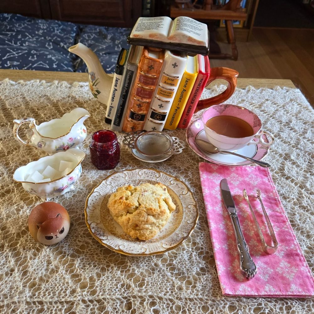 A teapot in the shape of all of Jane Austen's books sits on a lace covered table. The books are standing with their spines facing the camera. The teapot cover is an open book with the first few lines of Pride and Prejudice. The teapot is sitting on Orenburg lace. In front of the pot is a cup strainer and a small jar of raspberry jam. In front of that is a gold trimmed plate with a scone sitting on it. To the left side is a creamer and sugarbowl. They are decorated with flowers and they are gold trimmed. In front of those is a cat teapet. To the left side is a pink and creme vintage teacup. It is also gold trimmed. On the saucer of the cup is a silver spoon. In front of the cup is a pink and white cloth napkin. On top of it is a small silver butter knife and silver sugarcube tongs.