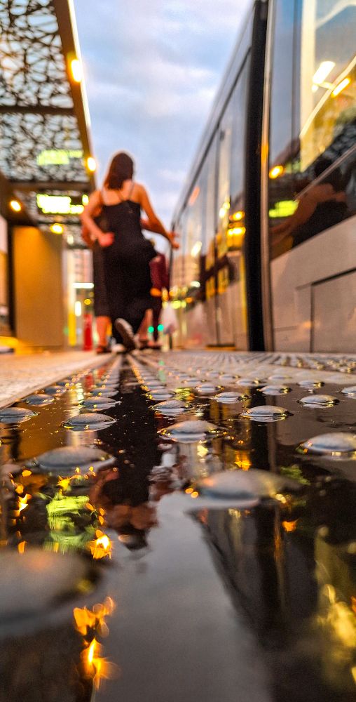 Photo du tram de Liège, à l'arrêt Feronstrée. Les lumières du soir se reflètent dans les flaques laissées par la pluie 