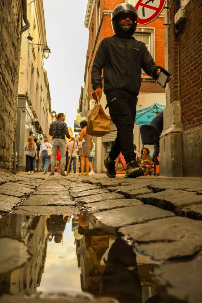 Photo de la rue du Diamant à Liège. Prise de vue au niveau du sol, près des pavés et d'une flaque qui reflète l'image. Des gens se promènent dans le Carré, un livreur avec un casque apparaît au premier plan 