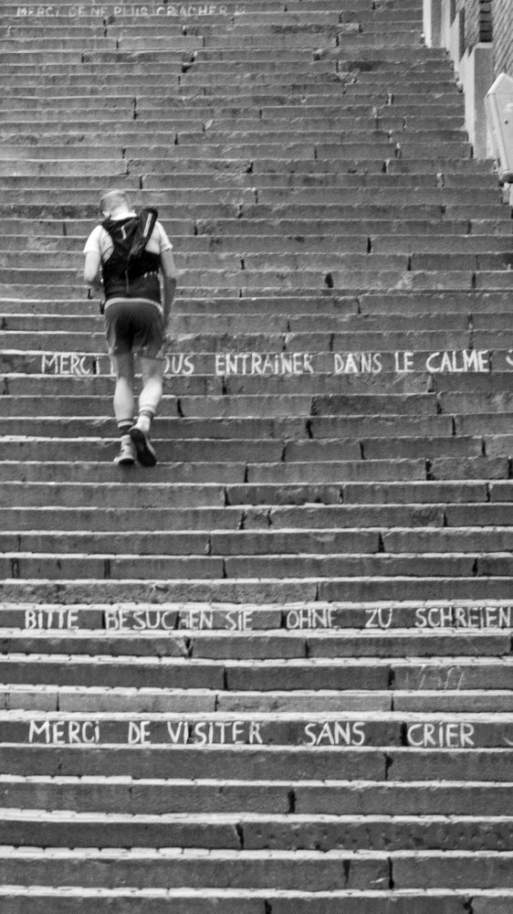 Photo en noir et blanc de la Montagne de Bueren à Liège, un joggeur grimpe, sur les marches on peut lire les inscriptions "merci de vous entraîner dans le calme" et "merci de visiter sans crier"