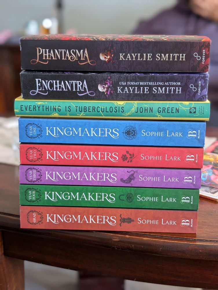 A stack of books on a dark wood coffee table.

Top to bottom: Phantasma and Enchantra by Kaylie Smith, Everything is Tuberculosis by John Green, and the Kingmakers series by Sophie Lark.