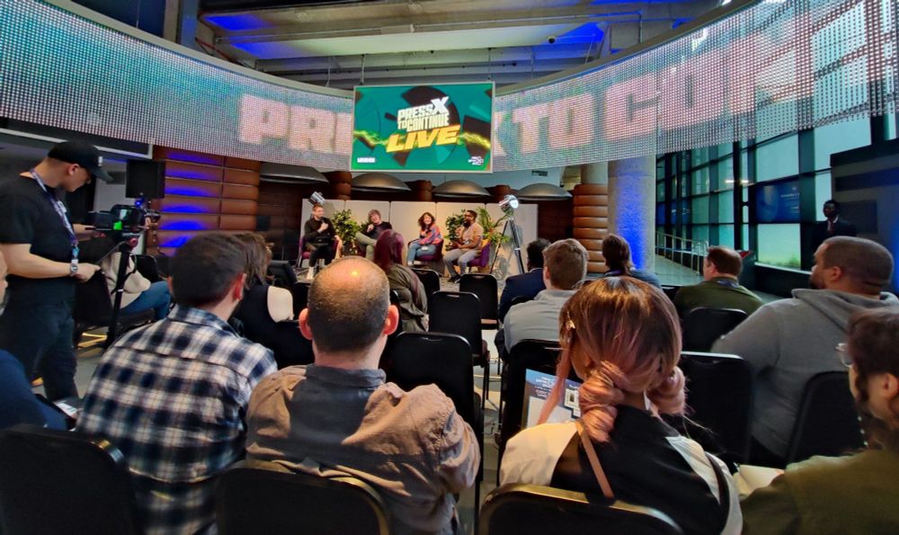 The cast of Press X To Continue on a stage, with purple chairs flanked by green foliage, interacting with the audience Live at London Games Festival. 