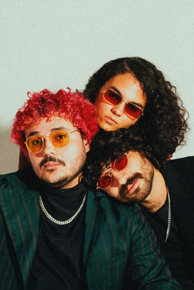 três jovens. um (eu) de cabelo rosa, óculos laranja e blazer verde, no meu ombro gabriel de óculos vermelho, bigode, blazer preto e colar de prata, apoiada na cabeça de gabriel, catarina, cabelo preto longo, óculos vermelho, cara de séria