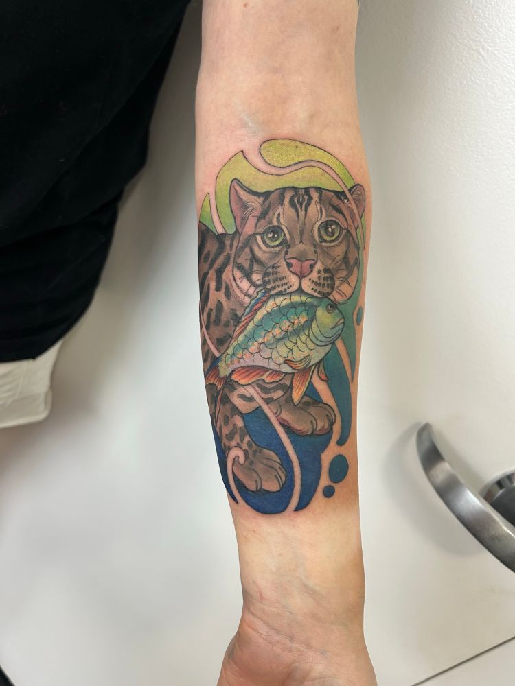 Inner forearm tattoo of a fishing cat holding a fish surrounded by a watery background