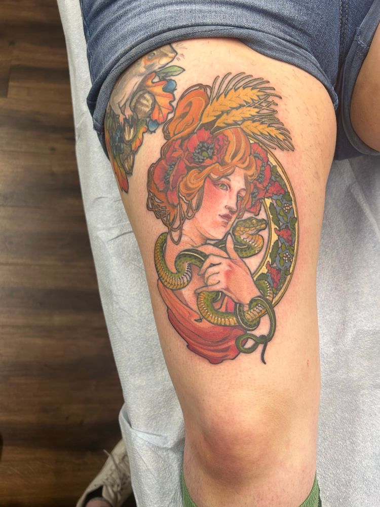 Art nouveau style thigh tattoo of Greek goddess Demeter surrounded by her symbols- snake, grapes, harvest wheat, poppies- in shades of red and rust and olive.