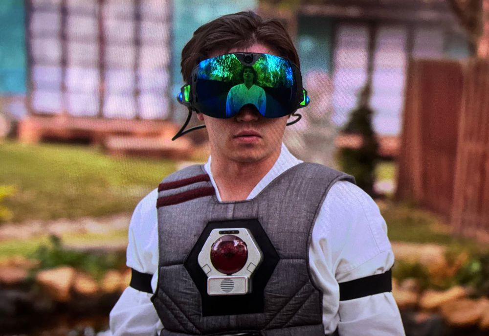 Still frame image of a character in Cobra Kai wearing a Lazer Tag sensor on his chest, along with virtual reality goggles 