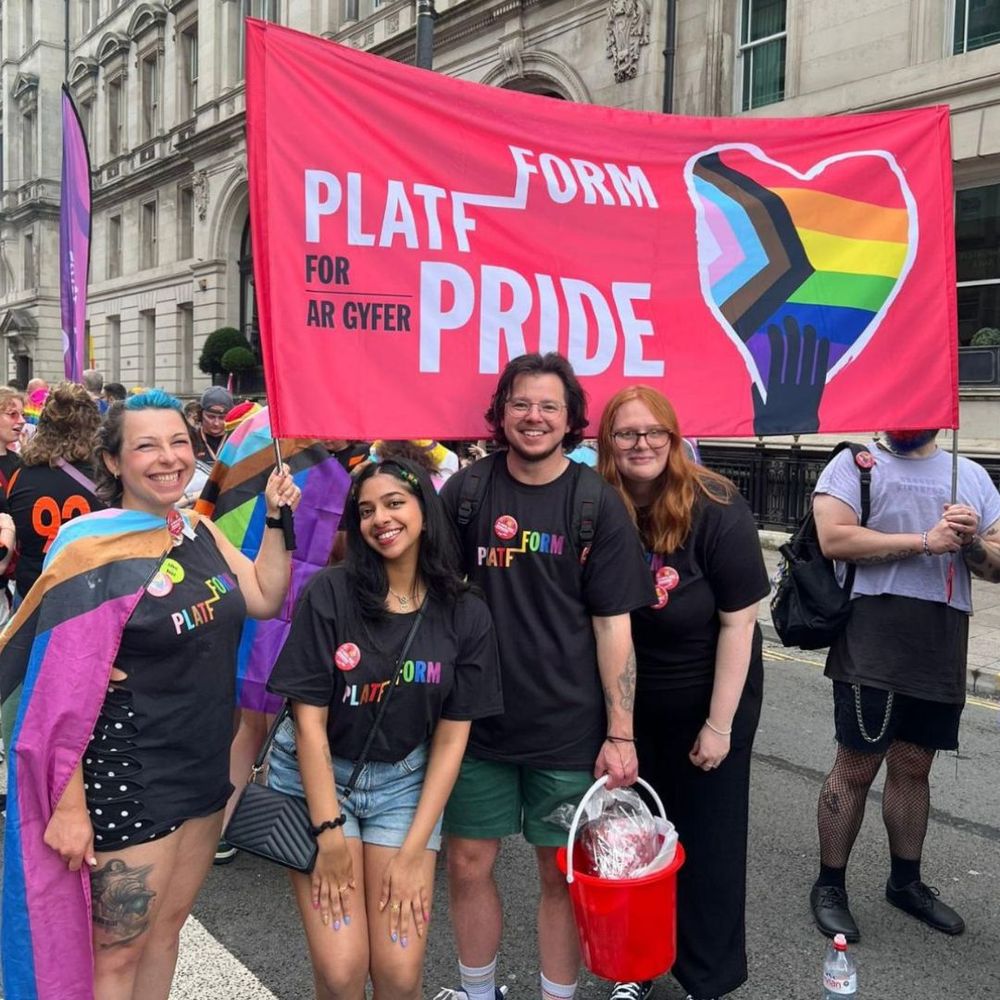 A group of smiling people stand in a street in Cardiff holding a banner reading 'Platfform for Pride / Platfform ar gyfer Pride.'