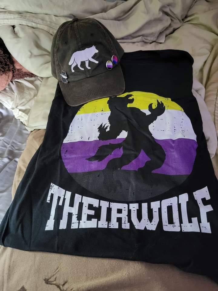 black t-shirt with a nonbinary flag in a circle with the outline of a werewolf and the text "THEIRWOLF"