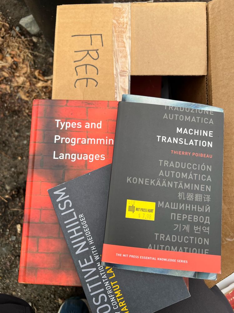 Picture of three books stacked on top of a cardboard box labeled "free." Books are Machine Translation by Thierry Poibeau, Positive Nihilism: My Confrontation with Heidegger by Hartmut Lange, and Types and Programming Languages