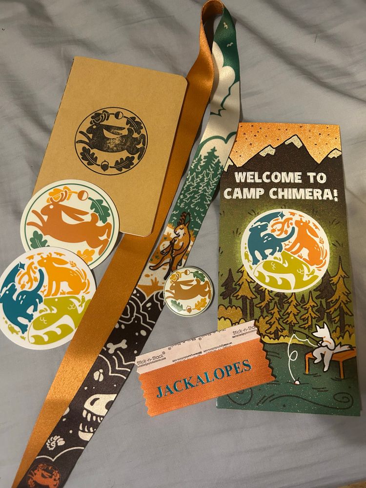 From top left to bottom right there is a small journal, two stickers, a lanyard, a pin, a jackalope name tag, and a Welcome To Camp Chimera! pamphlet. The journal, sticker, and pin have a jackalope emblem, with the other sticker and logo on the pamphlet having an orange Jackalope, blue opossum, and green tadpole dragon. The lanyard has a Jackalope furry in the middle, with trees behind it, and a soil layer having bones. The pamphlet background is of a furry fishing in a river, as they sit on a dock, with a forest behind them. 