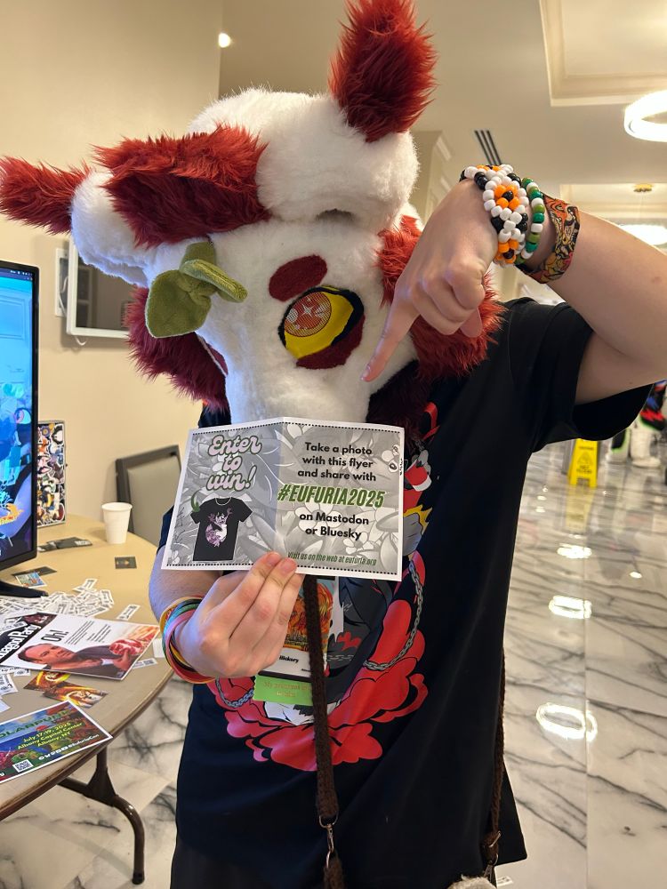 Me holding up a flyer while pointing down at it, wearing my red and white binturong fursuit head with a sprout on it