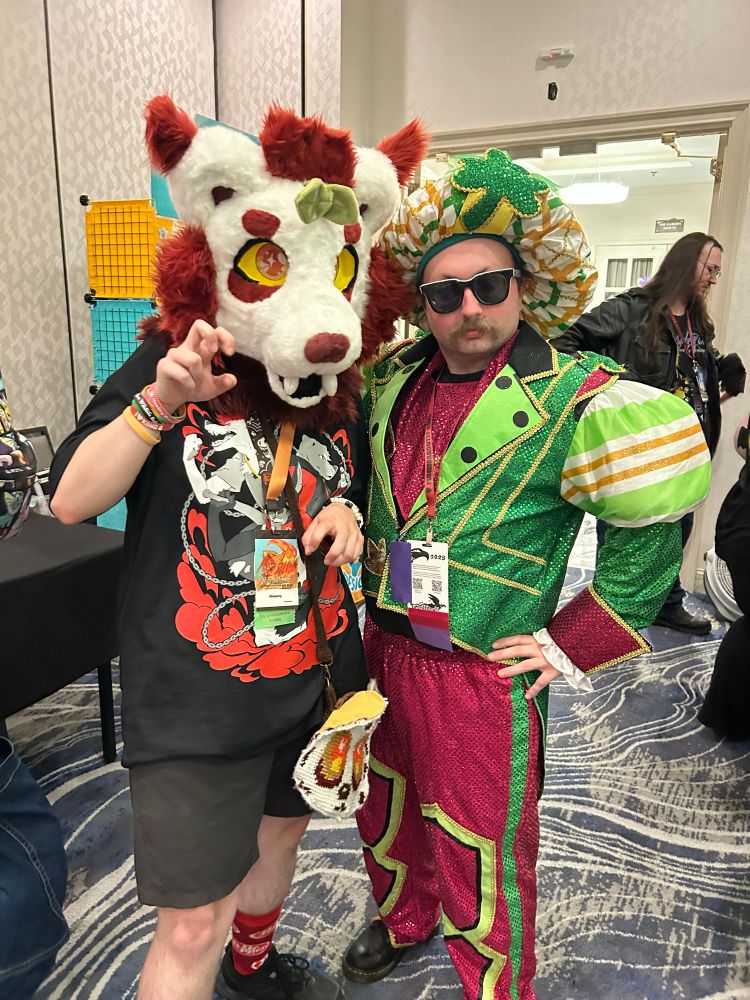 A red and white binturong fursuit posing with a very nicely dressed man in a red and green outfit