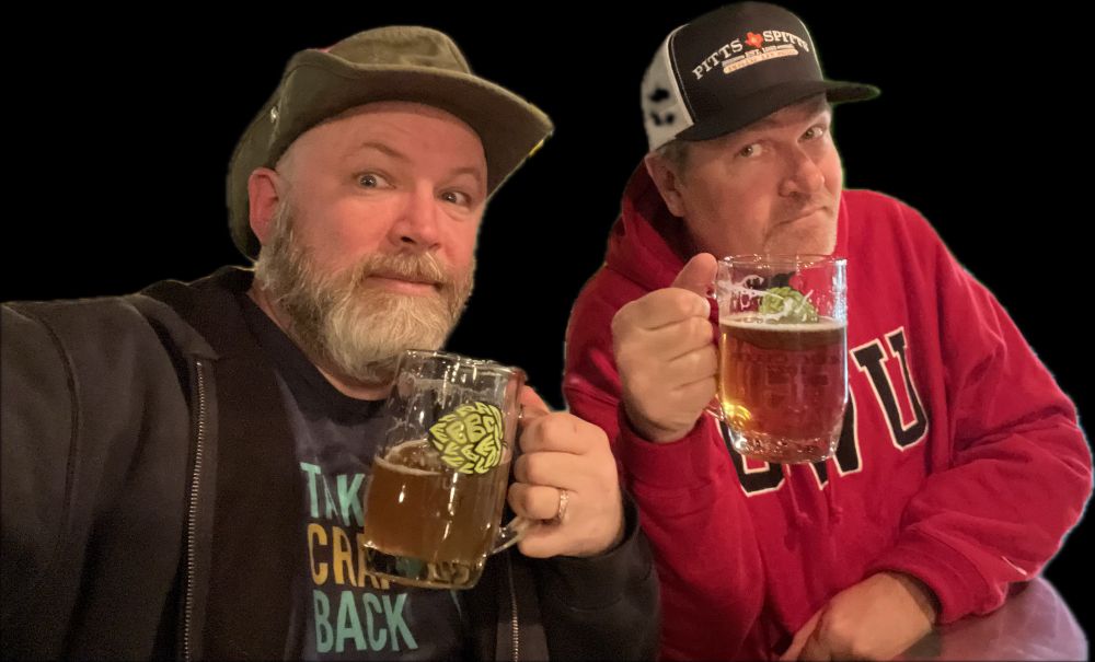 two guys holding beer mugs doing a cheers to the camera one wearing a green hat, one wearing a ball cap.