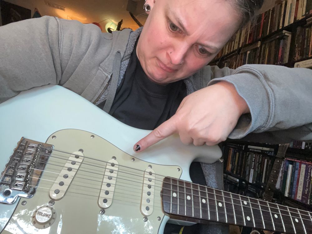 A woman holds a sonic blue Fender player’s strat while pointing at the middle pickup and jokingly making a silly face.