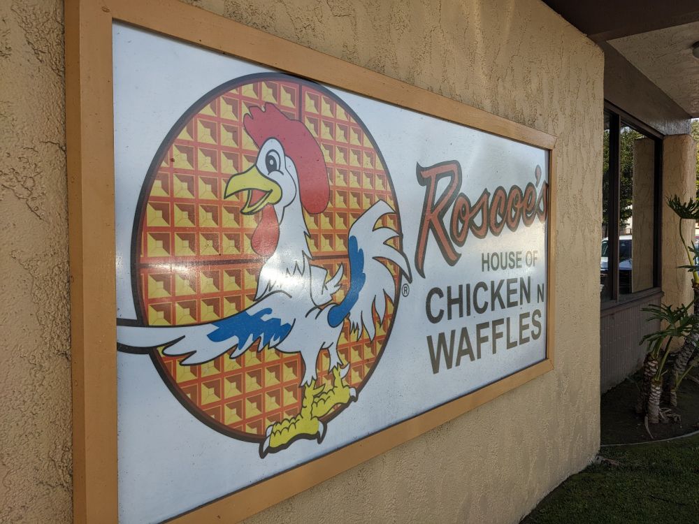 A large sign affixed to the side of a tan building. The sign says "Roscoe's House of Chicken and Waffles" next to picture of a smiling cartoon chicken in front of a background of a round waffle.
