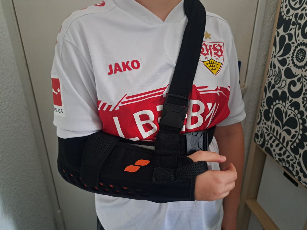 Das Foto zeigt eine junge Person, die ein weißes Fußballtrikot des VfB Stuttgart trägt. Das Trikot hat rote Akzente und den JAKO-Schriftzug.
​Die Person trägt am rechten Arm eine schwarze Armschlinge zur Ruhigstellung. Der Unterarm und Ellbogen sind in der schwarzen Stütze fixiert. Eine breite schwarze Gurt führt über die linke Schulter und hält den Arm in einer angehobenen Position vor dem Körper. Das Ende des rechten Arms (die Hand) ist sichtbar und greift leicht an den Bauchbereich.