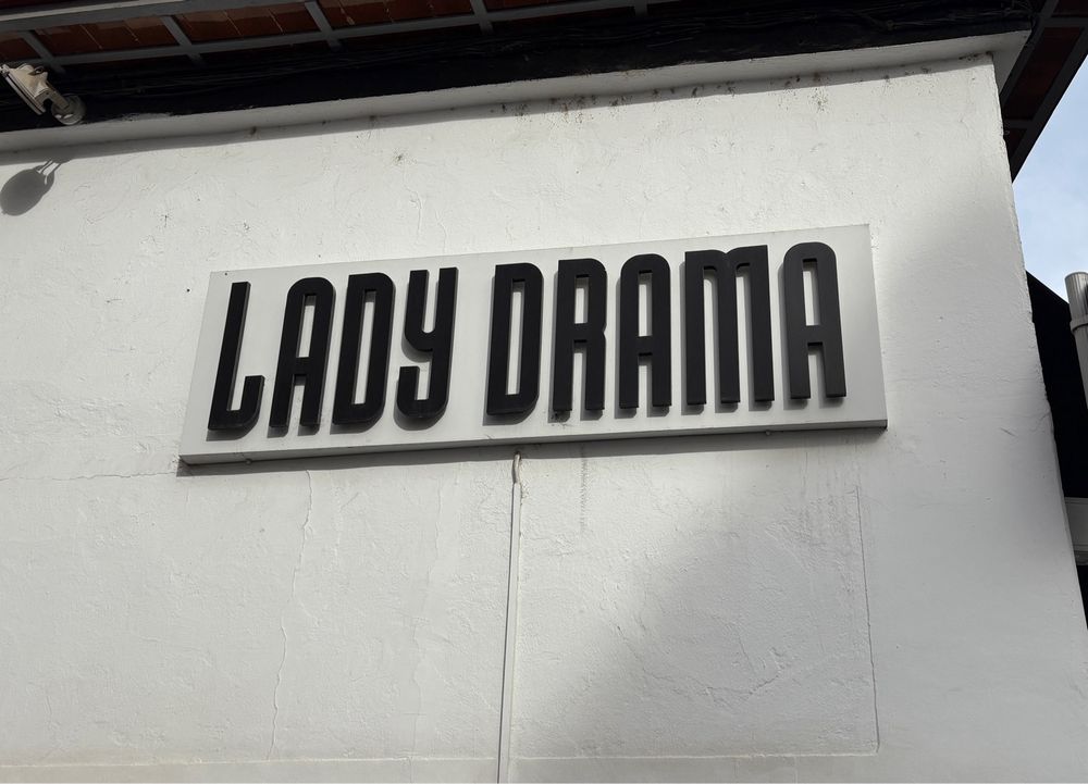 a black sign on a white wall, with long letters in all capitals, saying ‘LADY DRAMA’.