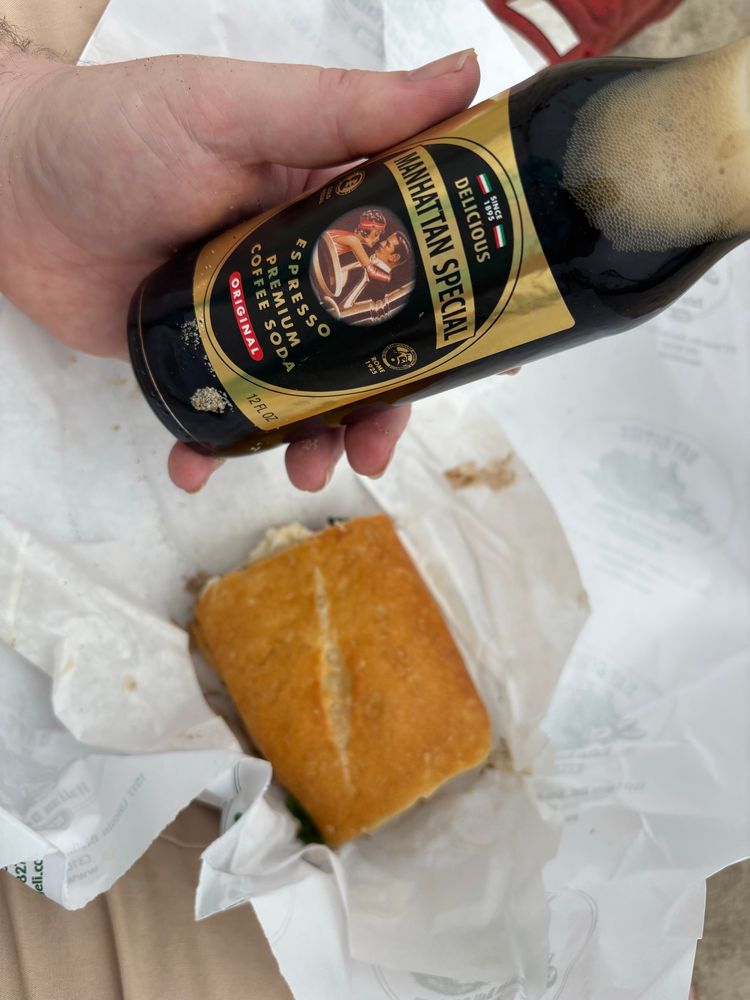 Photo of a birthday lunch: left hand holding a “Manhattan special” which is an espresso premium coffee soda, and an unwrapped burrata caprese sandwich from Bay Cities Italian deli. 