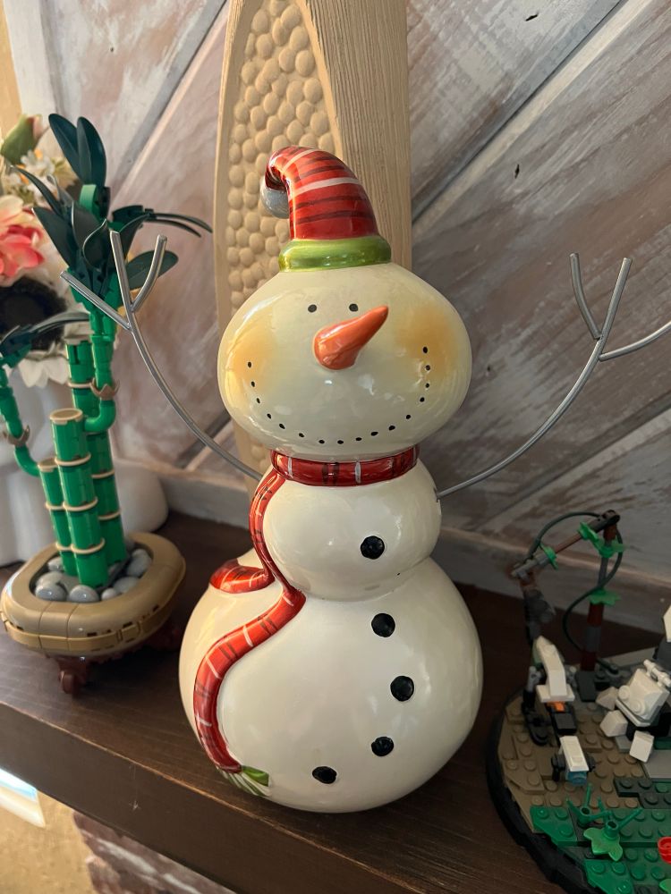 Cute white ceramic snowman wearing a jaunty knit cap. He has a carrot for a nose & a red ribbon accent around his neck. 