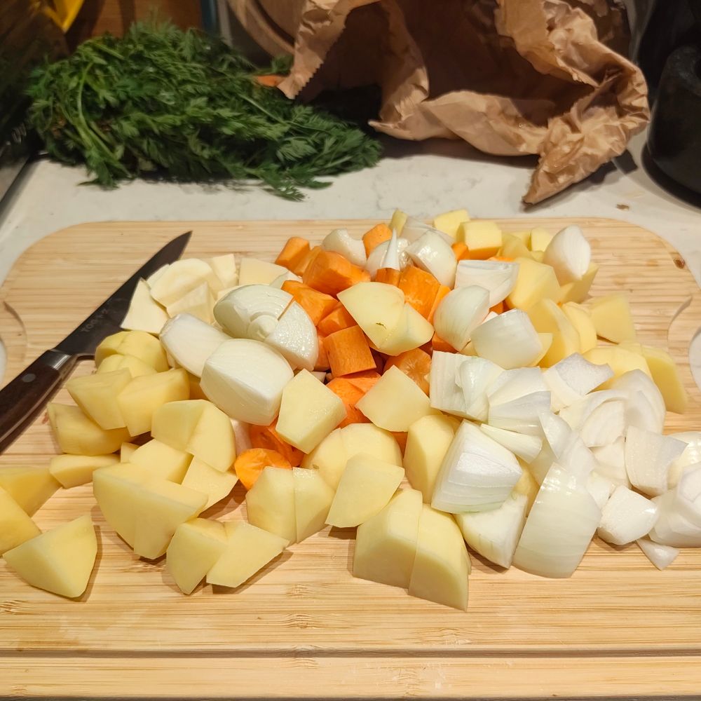 Roughly chopped potato, carrots, parsnips and onions on a wooden board with a knife next to them. All of it, and the beef out of shot, was picked up from a weekly farmers market just round the corner where everything is from local supplier which makes this actually a feel a little bit more of a special first time with the knife. 