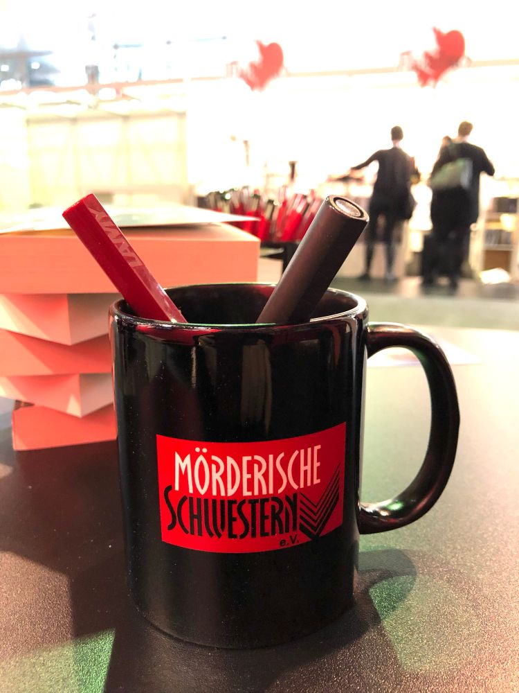 Eine Tasse des Krimiautorinnenvereins „Mörderische Schwestern“ auf der Frankfurter Buchmesse. Darin in den Vereinsfarben ein schwarzer und ein roter Füller, im Hintergrund ein Bücherstapel und schemenhaft Messebesucher.