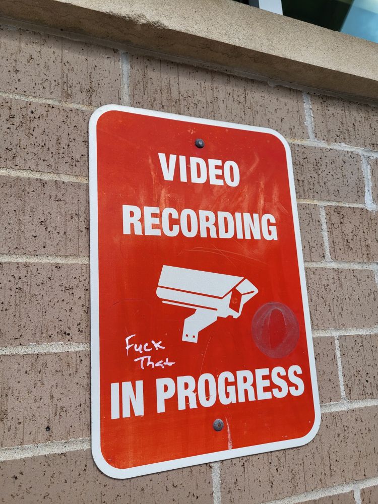 A red sign that says "video recording in progress," on which someone has written "fuck that"