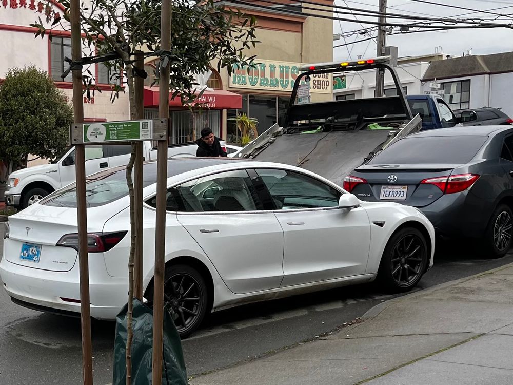 White Tesla sedan parked crookedly, directly in a residential driveway. Flatbed tow truck is visible behind it with its bed inclined to load the vehicle onto the bed.