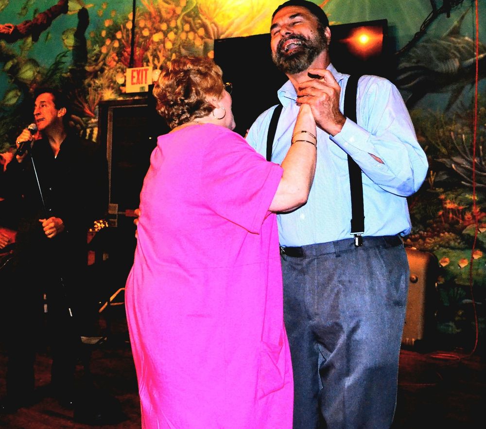 The image depicts a man and a woman dancing in a dimly lit venue. The woman, viewed from behind, is wearing a bright pink dress and is holding hands with the man, who is facing her and smiling with his eyes closed. He is wearing a light blue button-down shirt, grey pants, and black suspenders. In the background, to the left, another man is singing into a microphone. An "EXIT" sign is visible above the stage area. The background features a colorful mural backdrop with undersea images.
