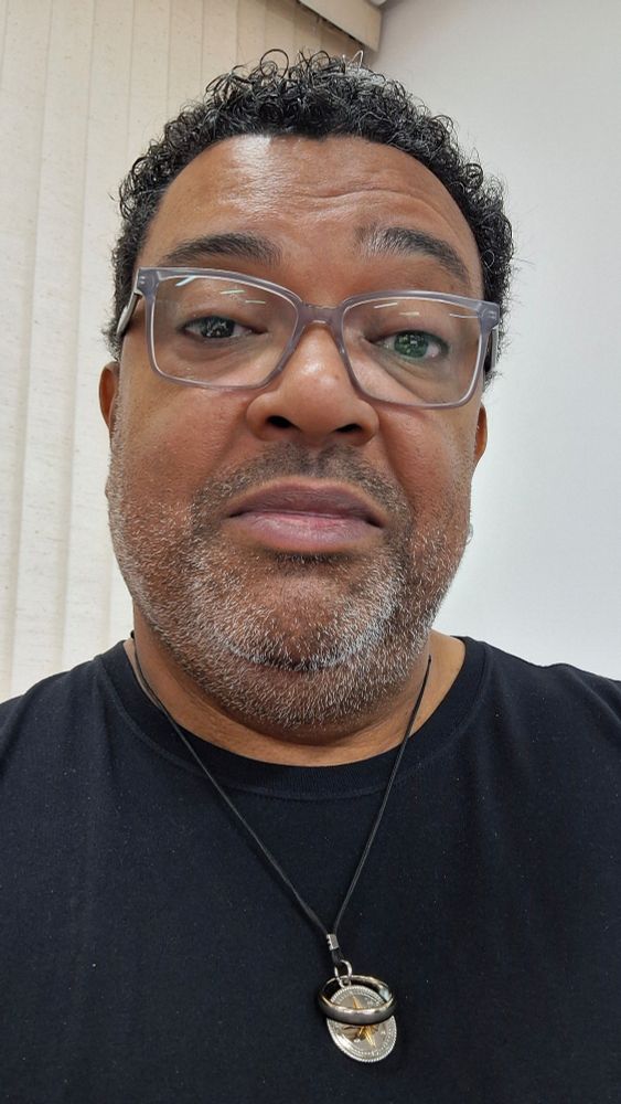 Eu de camiseta preta, óculos de grau, barba por fazer e colar com rosa dos ventos e minha aliança de casamento. 