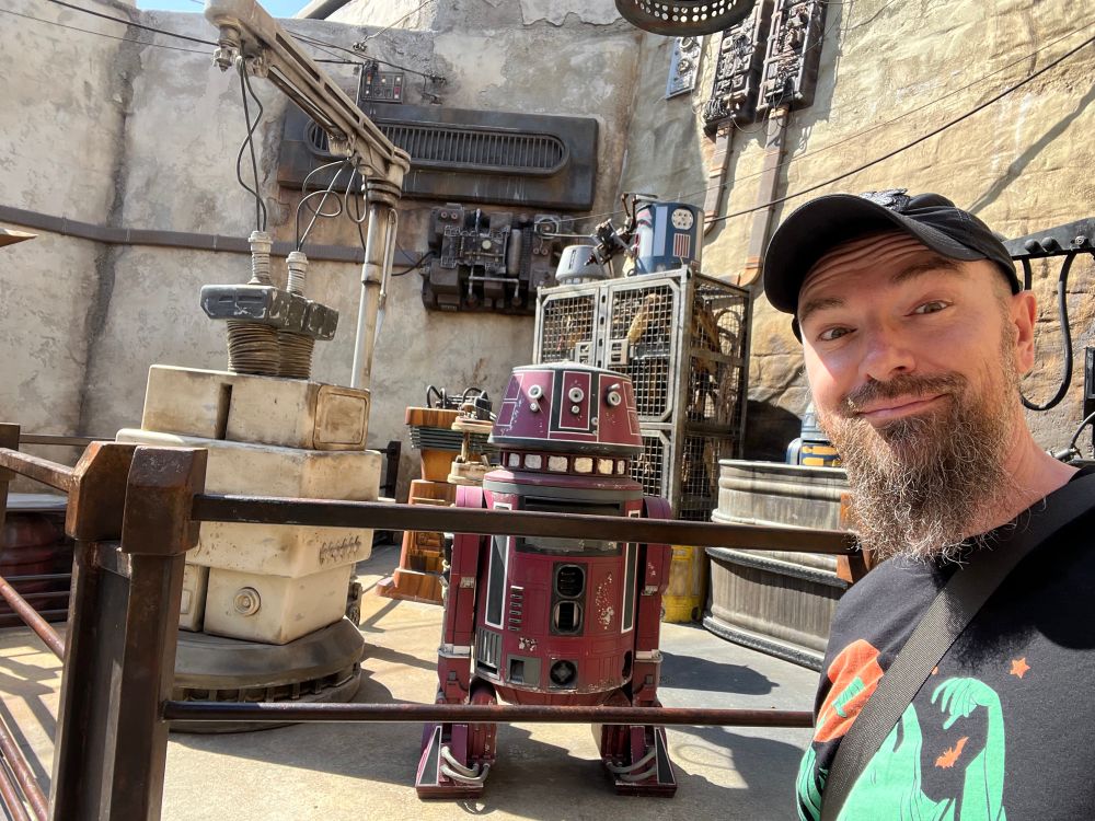 A handsome bearded man making an amused face next to a Star Wars droid 